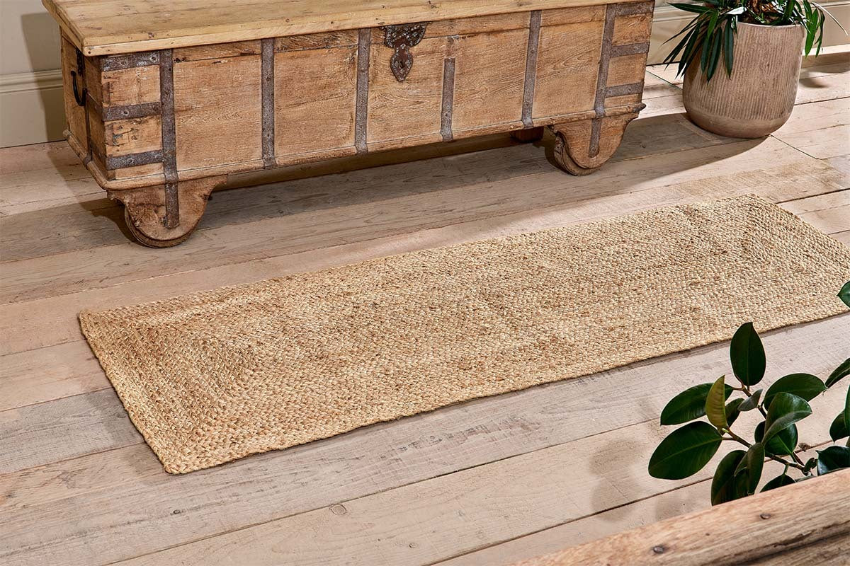 The nkuku Braided Hemp Floor Runner, a rectangular eco-friendly piece, lies on a light wooden floor by a rustic chest. Green potted plants at the edges enhance the cozy, natural look.