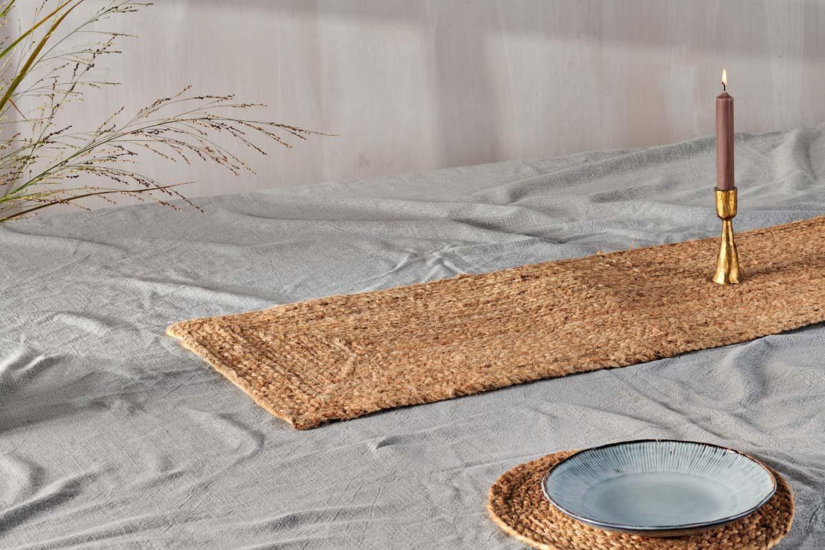 A light gray cloth-covered table features artisan tableware, a brass candle holder with a mauve candle, and the nkuku Braided Hemp Table Runner with a matching placemat beneath a blue-gray ceramic plate. Dried grass accents the tablescape.