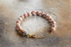 The Cerita Bracelet - Rhodochrosite by nkuku features round, marbled pink and white semi-precious beads, a gold chain extension, and a gold inhale tag, shown on a textured beige and brown background.