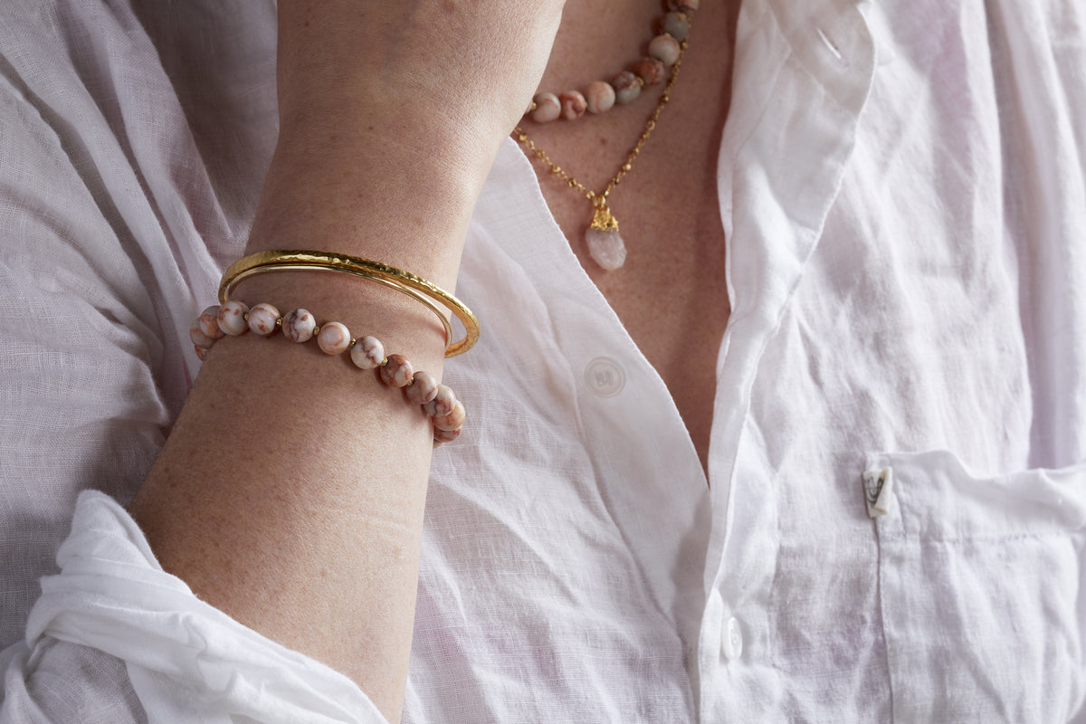 A person in a white open-collar shirt wears nkukus Cerita Bracelet—crafted from pink rhodochrosite—paired with a matching necklace, gold bangle, and gold pendant necklace for a versatile accessory collection.