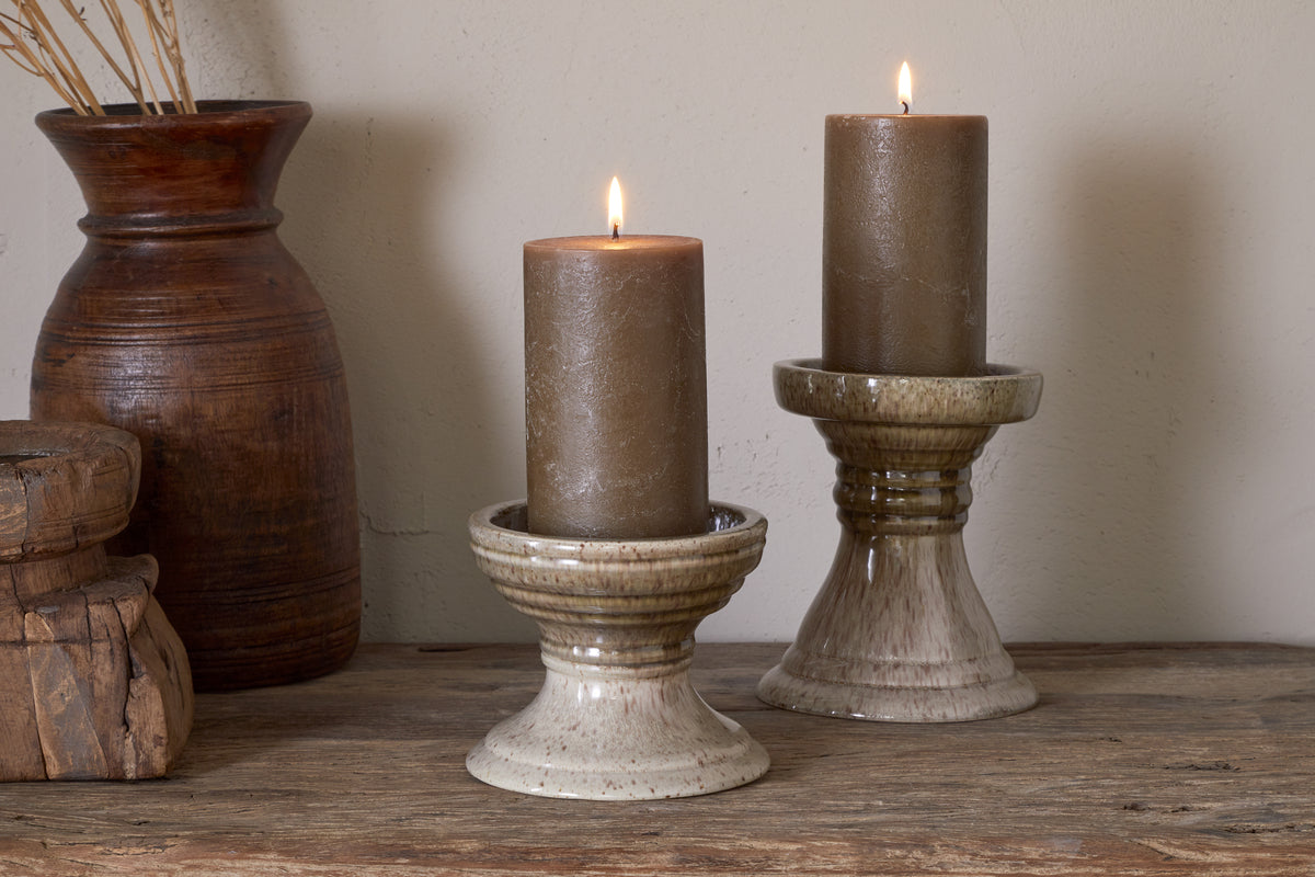 Two brown candles glow on nkuku’s Cabra Reactive Glaze Candle Holders in Olive & Natural, each at a different height. Set atop rustic wood with a wooden vase of dried grasses, they bring cozy, earthy style to a neutral wall.