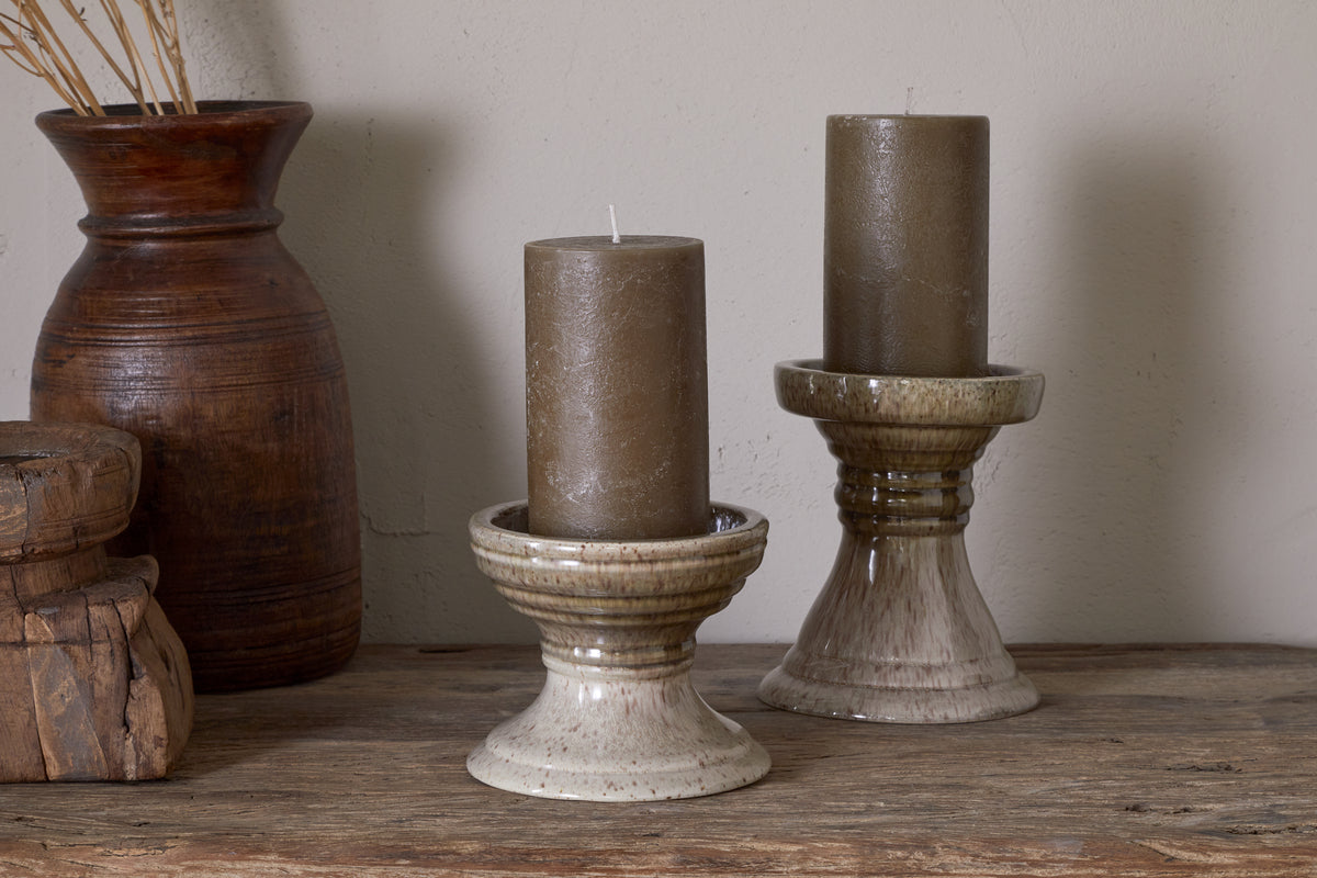 Two brown pillar candles sit in nkuku Cabra Reactive Glaze Candle Holders in Olive & Natural on a wooden surface, with a tall brown clay vase of dried grass and stacked wooden boxes creating an earthy, rustic vintage decor.