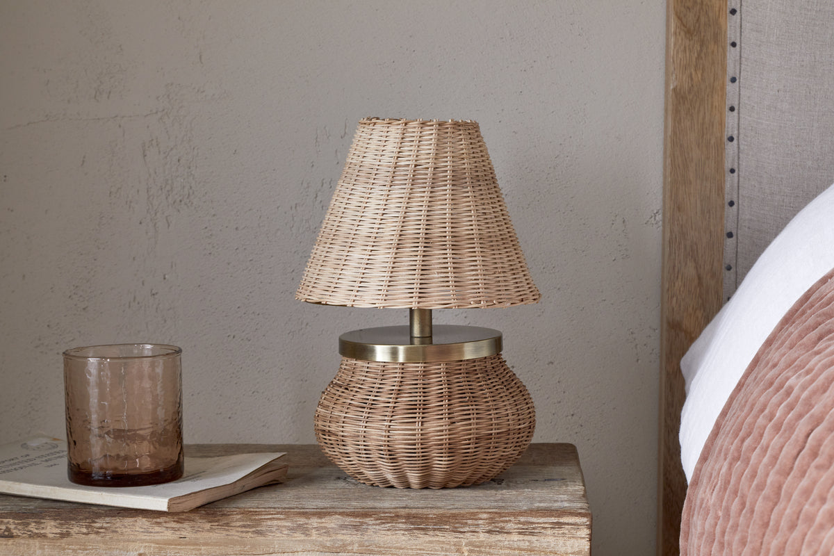 The nkuku Cija Rattan Table Lamp - Natural, with a woven shade and brass accent, sits on a rustic nightstand beside a brown glass tumbler, a closed book, and a bed with a pink pillow.
