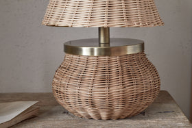 The Cija Rattan Table Lamp - Natural by nkuku, featuring a wide rounded base and matching rattan shade with brass accents, rests on a wooden surface beside a closed book.
