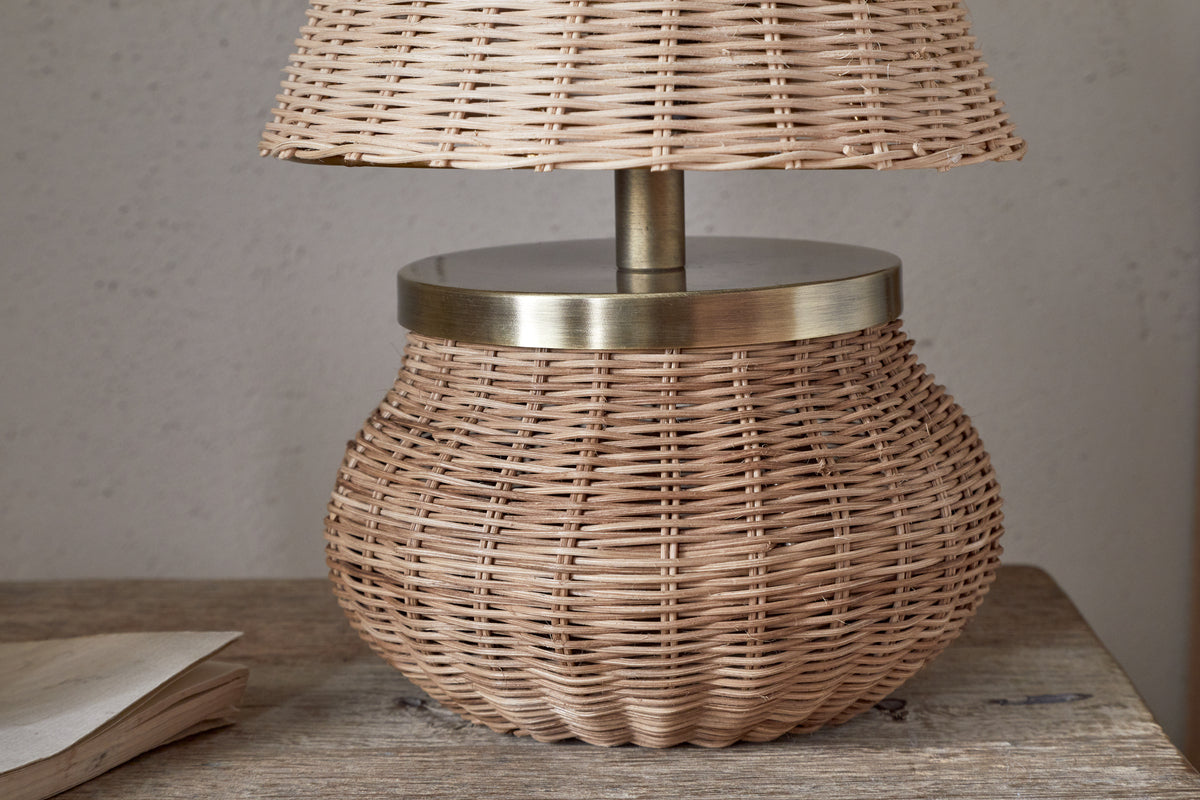 The Cija Rattan Table Lamp - Natural by nkuku, featuring a wide rounded base and matching rattan shade with brass accents, rests on a wooden surface beside a closed book.