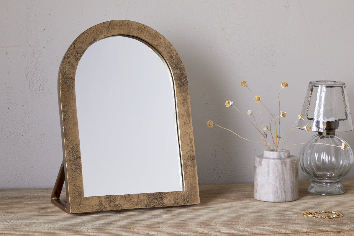 The nkuku Cayce Cast Aluminum Vanity Mirror in Antique Brass sits on a wooden dressing table beside a marble vase with dried flowers, a clear glass oil lamp, and a gold bracelet against a light, textured wall.