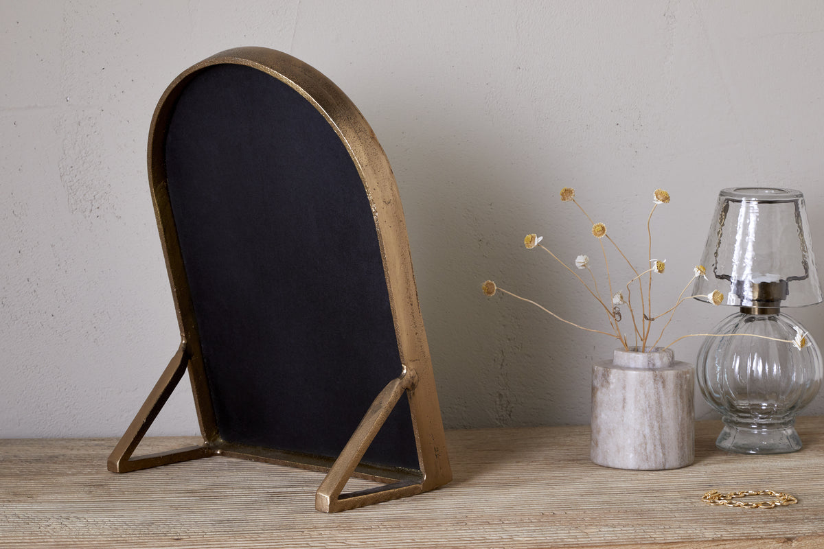 The nkuku Cayce Cast Aluminum Vanity Mirror in Antique Brass stands on a wooden dressing table beside a marble vase with dried flowers, a clear glass oil lamp, and a gold bracelet. The background features a neutral, plain wall.