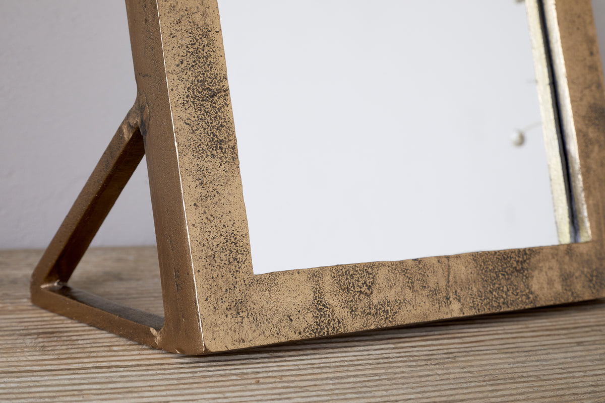 Close-up of the nkuku Cayce Cast Aluminum Vanity Mirror in Antique Brass, featuring a rectangular, textured frame resting on a light wooden surface. Ideal as a dressing table or standing vanity mirror.