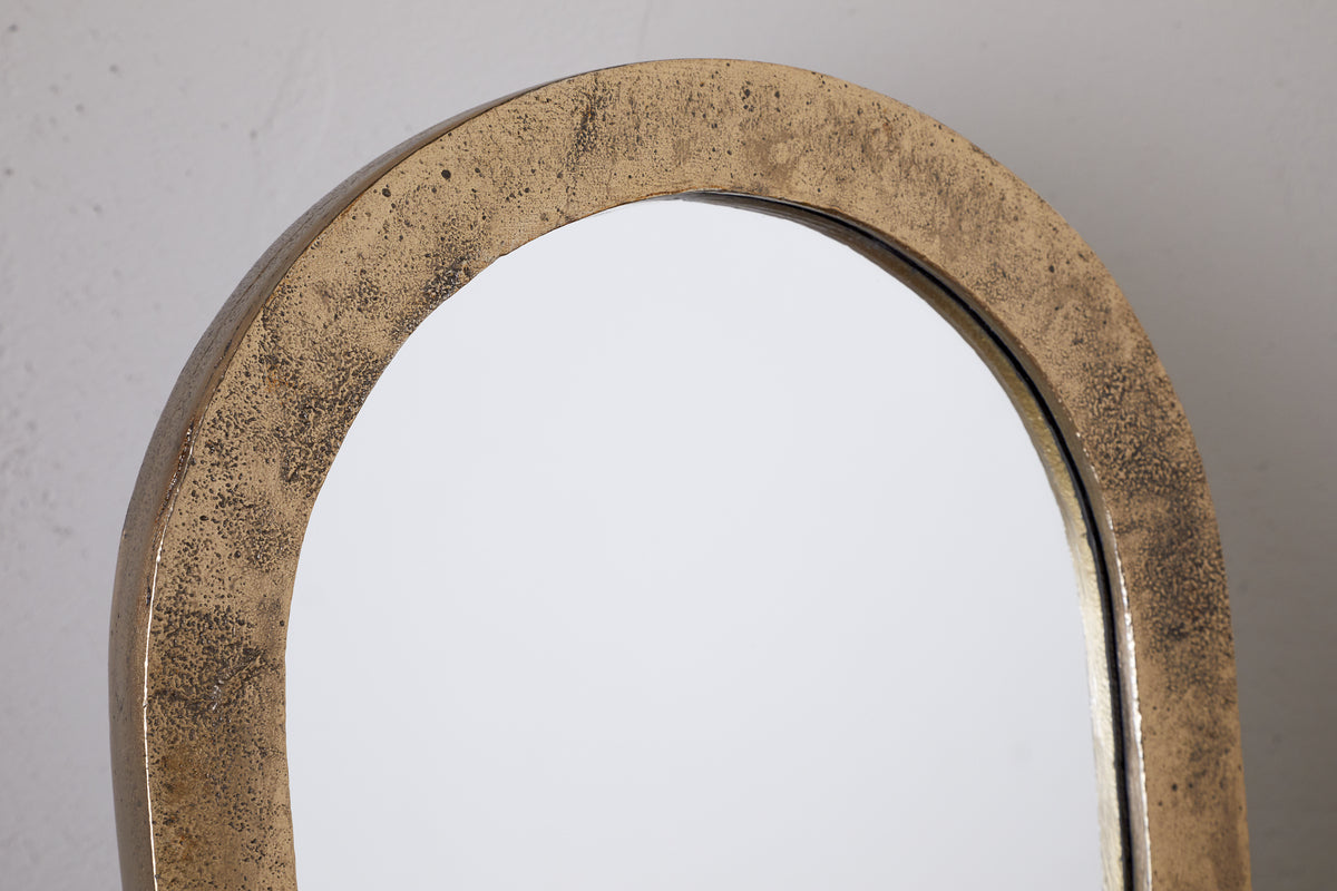 A close-up of the Cayce Cast Aluminum Vanity Mirror by nkuku, featuring an arched top and textured antique brass frame, set against a plain light background.