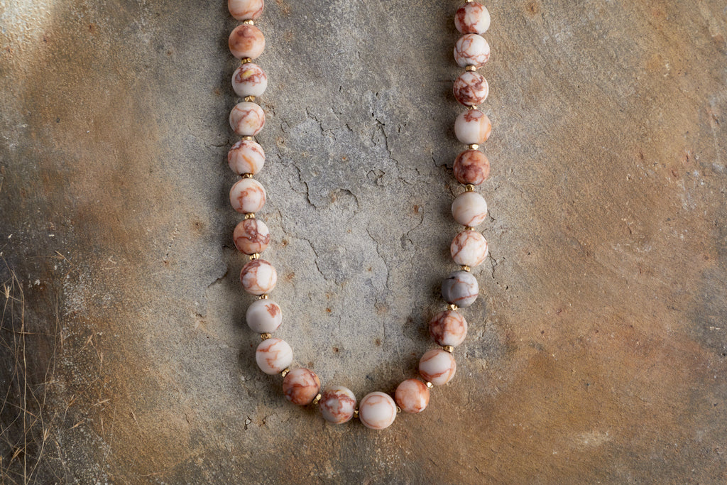 The Cerita Necklace - Rhodochrosite by nkuku features round, marbled pink and white semi-precious stones, beautifully displayed on a textured earthy brown surface.