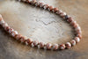The Cerita Necklace - Rhodochrosite by nkuku features round, marbled pink and white semi-precious stones, displayed on a rustic brown and beige surface—an elegant and versatile accessory for any occasion.