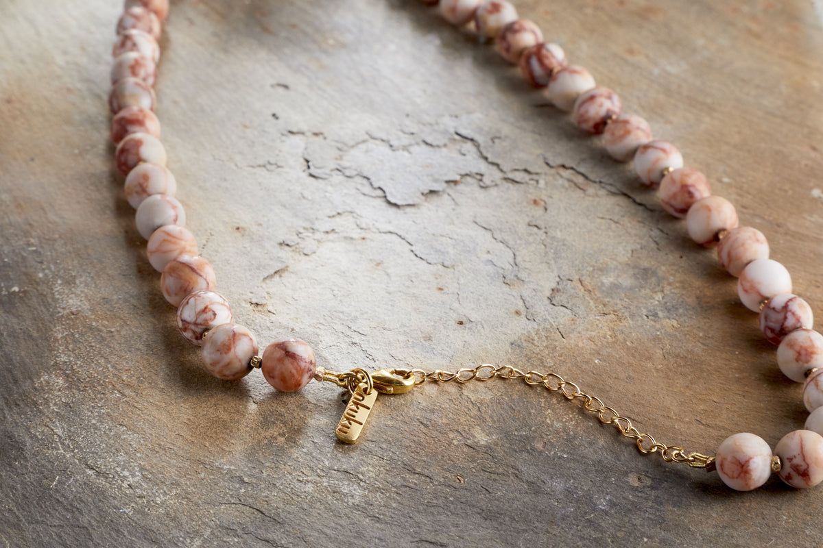 The Cerita Necklace - Rhodochrosite by nkuku features marbled pink and white semi-precious stones and a gold chain extender, displayed on an earthy textured surface—a versatile accessory for any occasion.