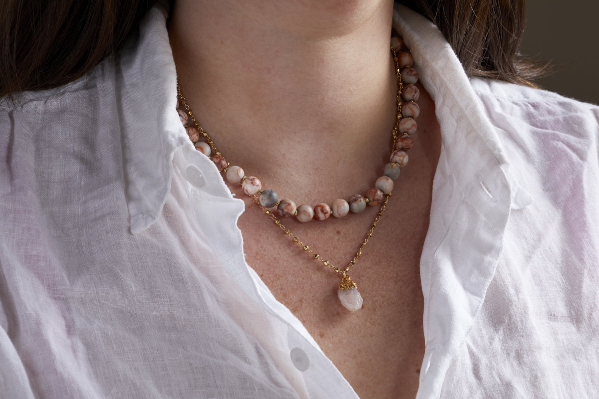 A woman in a white button-up shirt wears two layered necklaces, including nkukus Cerita Necklace - Rhodochrosite: a gold chain with a pink rhodochrosite pendant, paired with semi-precious stone beads for an effortlessly versatile look.