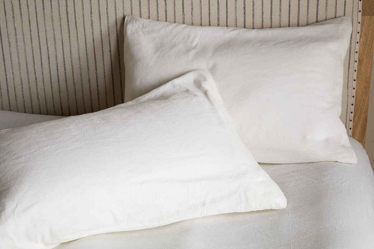 Two Cela Hemp & Cotton Housewife Pillowcases by nkuku in natural shade rest on a light bed with a striped headboard. One pillow stands upright, the other lies flat, suiting different bedroom styles with their breathable hemp and cotton blend.