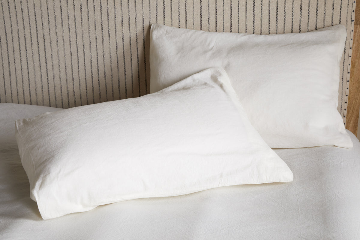 Two Cela Hemp & Cotton Housewife Pillowcases in natural by nkuku rest on a neatly made bed with a light, vertically striped headboard, creating a clean and minimalistic bedroom style.