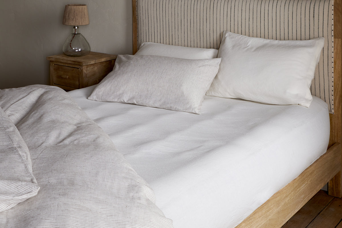A neatly made bed with nkukus Cela Hemp & Cotton Fitted Sheet in natural, two large pillows and one small pillow, a light upholstered headboard with vertical stripes, beside a wooden nightstand and glass lamp.