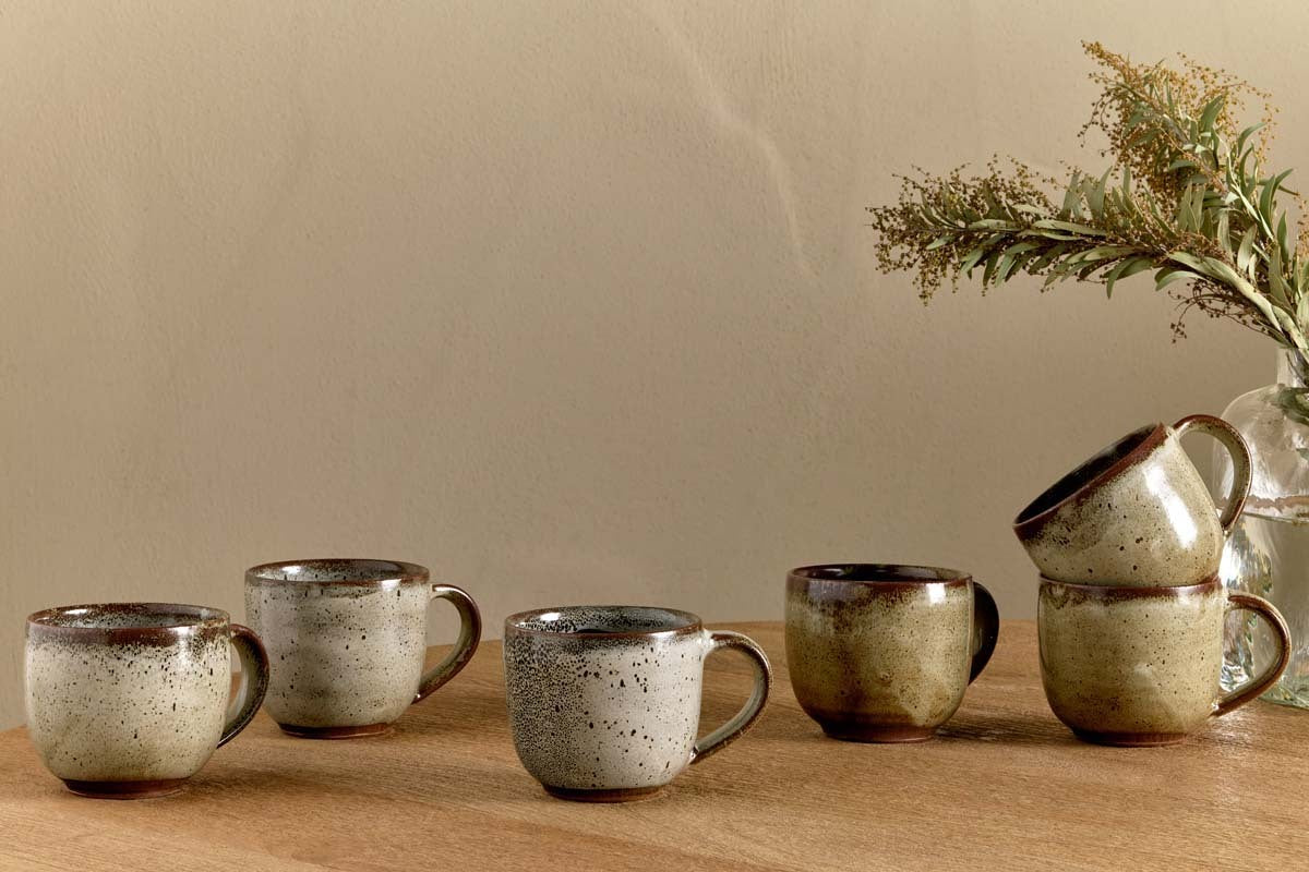Five nkuku Cam Coffee Mugs - Mocha (Set of 2) with reactive glaze are arranged on a wooden surface; three stand alone, two are stacked. A glass vase with dried greenery is set in the background against a beige wall.