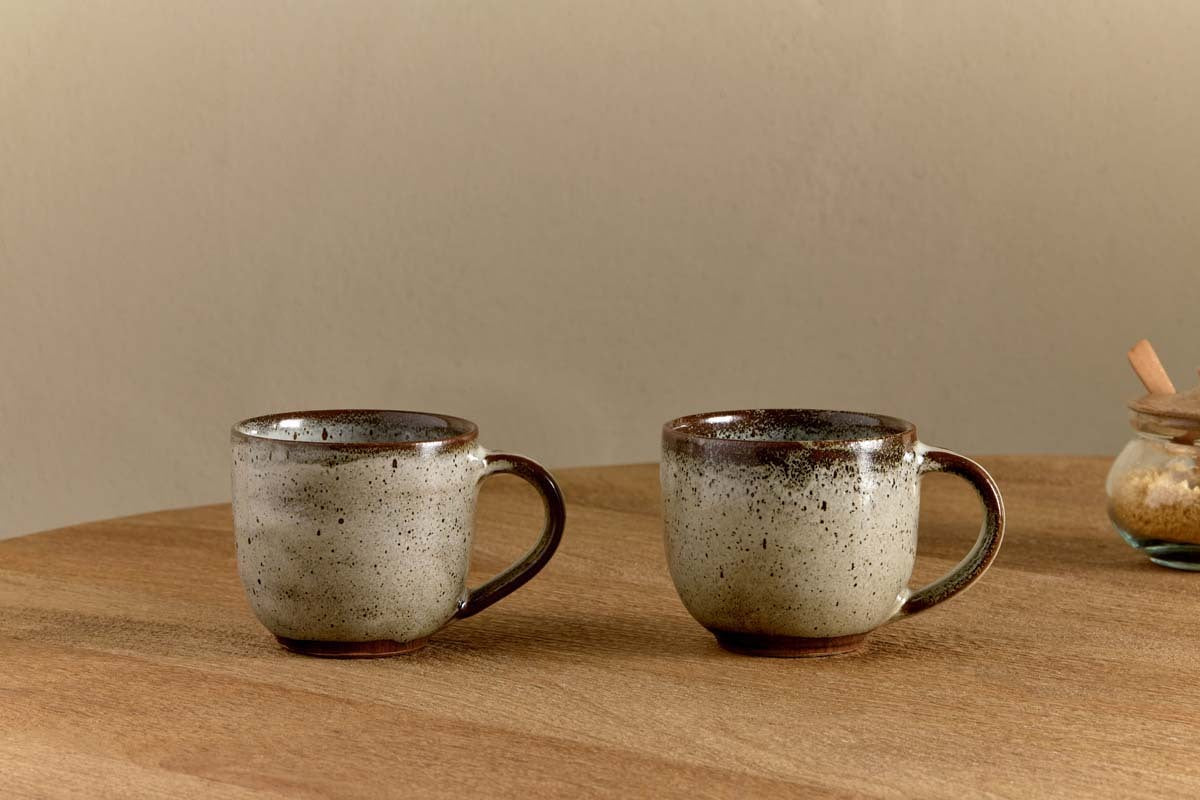 Two Cam Coffee Mugs - Mocha by nkuku (set of 2) with a speckled reactive glaze rest on a wooden table. A glass jar with a wooden lid appears on the right, all set against a neutral beige wall backdrop.