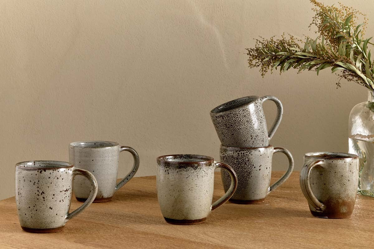 Six nkuku Cam Large Mugs in mocha, sold as a set of 2, feature earthy tones and reactive glaze. Arranged on wood with two mugs stacked, they sit beside a glass vase of eucalyptus against a neutral beige wall.