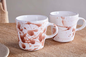 The nkuku Canela Mug set in Dusty Terracotta features two ceramic mugs with brown abstract splatter designs, offering a cozy, minimalist touch to your space. Perfect for enjoying beverages together or as a stylish gift.