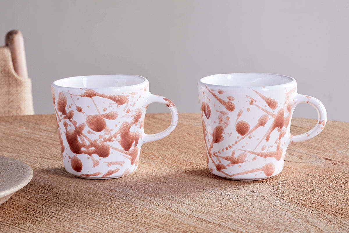 Two nkuku Canela Mug - Dusty Terracotta (Set of 2) feature white glaze with rust-hued splatter, round handles, and a tapered shape. They sit on a wooden table with linen napkins in the background.
