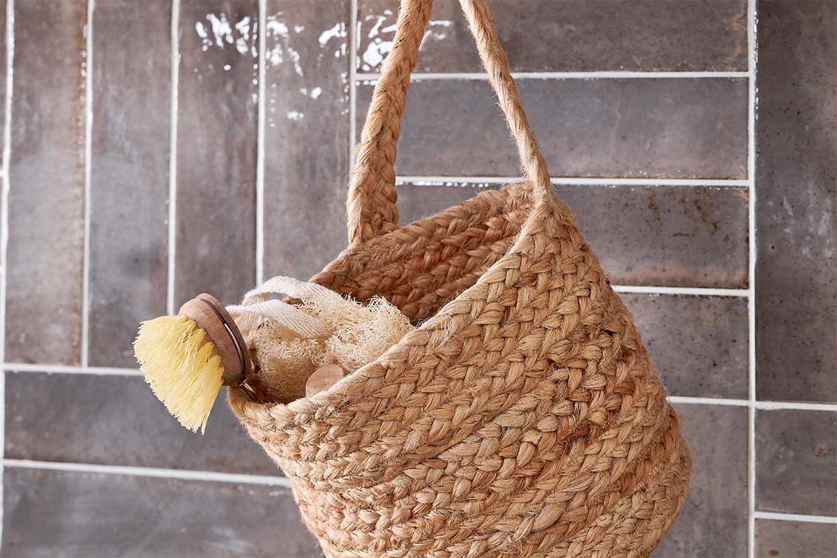 The nkuku Chapad Hemp Wide Wall Hung Basket hangs on glossy grey and white tiles. Inside, a wooden-handled scrub brush and loofah sponge hint at its use for stylish bathroom or kitchen storage.