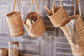 Four nkuku Chapad Hemp Wide Wall Hung Baskets hang on hooks against tiled walls, each holding utensils and a cloth. A glass jar with a wooden lid sits on the glossy gray tiles beneath the baskets.
