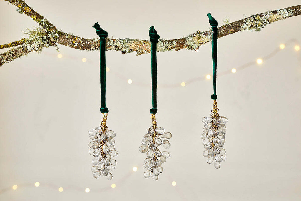 The Danvi Glass Drop Baubles - Brass & Clear (Set of 3) by nkuku hang from a tree branch, accented by twinkling string lights for a festive and elegant Christmas display.