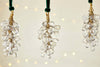 The nkuku Danvi Glass Drop Baubles - Brass & Clear (Set of 3) feature clusters of clear glass beads wrapped in brass wire and hang from a velvet tie, glowing with warm lights for a festive Christmas tree ambiance.