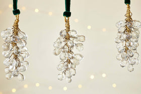 The nkuku Danvi Glass Drop Baubles - Brass & Clear (Set of 3) feature clusters of clear glass beads wrapped in brass wire and hang from a velvet tie, glowing with warm lights for a festive Christmas tree ambiance.
