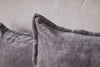 Two nkuku Dhara Velvet Cushion Covers in charcoal, featuring visible seams, rest against a light beige fabric backdrop for a soft and cozy look with an elegant touch.