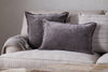 A light beige sofa styled with two large gray velvet pillows, nkukus Dhara Velvet Cushion Cover in Charcoal, a textured gray throw blanket, and a cream pillow with thin black stripes creates a cozy, modern living room vibe.