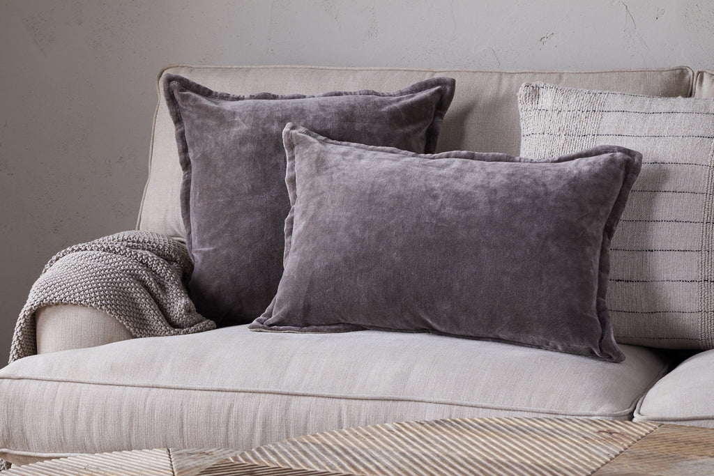 A light beige sofa styled with two large gray velvet pillows, nkukus Dhara Velvet Cushion Cover in Charcoal, a textured gray throw blanket, and a cream pillow with thin black stripes creates a cozy, modern living room vibe.
