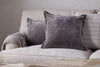 A beige sofa with two nkuku Dhara Velvet Cushion Covers in charcoal, a textured gray knit blanket over the arm, and a beige pillow with black stripes, all set against a light wall.
