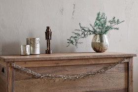 A rustic wooden chest features the nkuku Danvi Glass Drop Garland in brass and clear draped across the front. On top sit two metallic candle holders, a bronze figurine, and a silver vase with green foliage, set against a textured beige wall.
