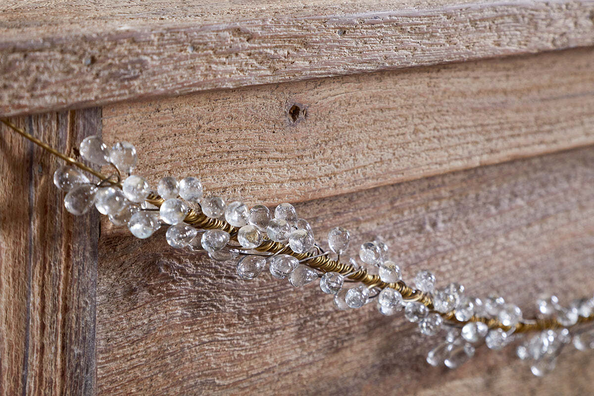 A close-up of a wooden surface decorated with the nkuku Danvi Glass Drop Garland in Brass & Clear, featuring twisted gold wire and crystal-like glass drops draped diagonally for a festive look.