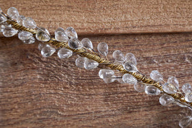 The Danvi Glass Drop Garland by nkuku features clear teardrop glass beads on brass wire, elegantly draped over a textured brown wooden surface, catching the light and adding a sparkling accent to festive table decor.