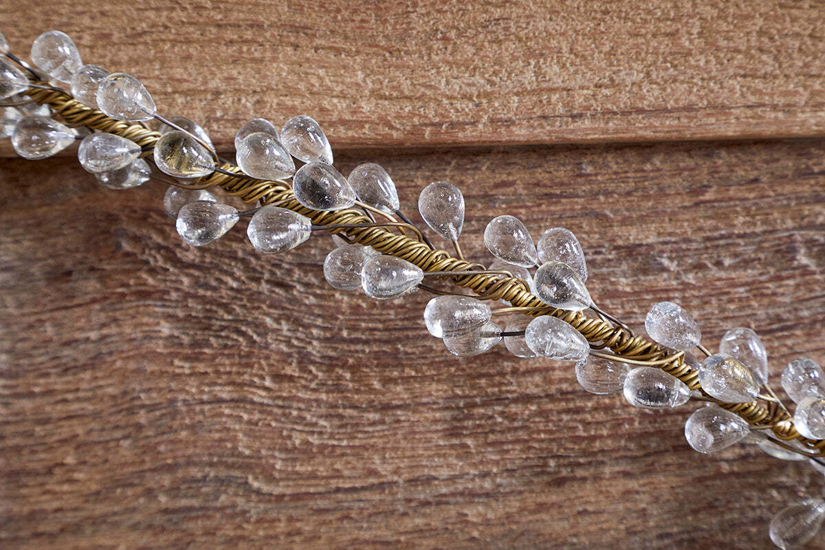 The Danvi Glass Drop Garland by nkuku features clear teardrop glass beads on brass wire, elegantly draped over a textured brown wooden surface, catching the light and adding a sparkling accent to festive table decor.