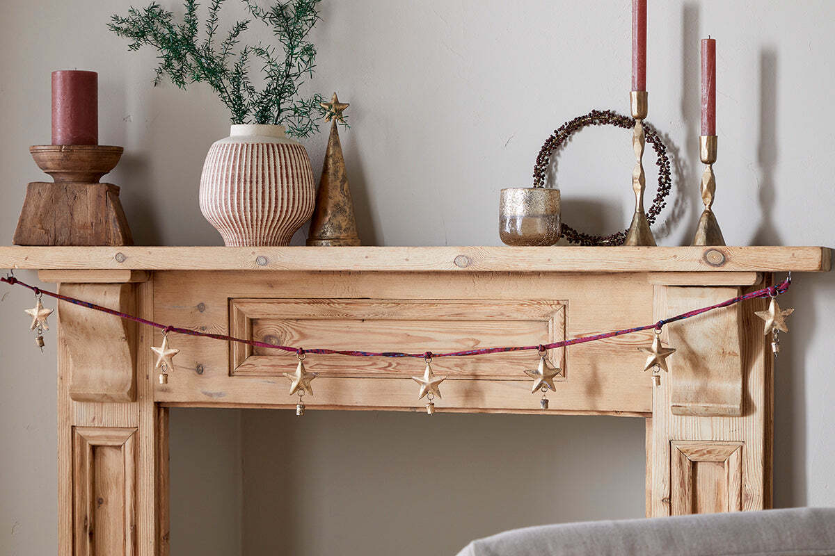 A rustic wooden mantel is decorated with nkuku’s Didi Star Garland in antique brass, two red candles, a pink-striped vase of greenery, a metallic cone tree, a round twig wreath, and a small metallic cup against a light beige wall.
