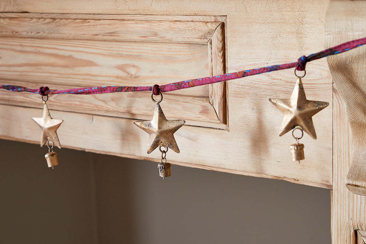 The nkuku Didi Star Garland features three handcrafted antique brass stars, each with a small key, hanging from a colorful ribbon and displayed on natural wood for a festive, artisanal touch.