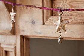 A close-up of the nkuku Didi Star Garland - Antique Brass, featuring antique brass stars and small bells on a twisted pink and red cord, draped over a rustic wooden cabinet with a softly blurred background.