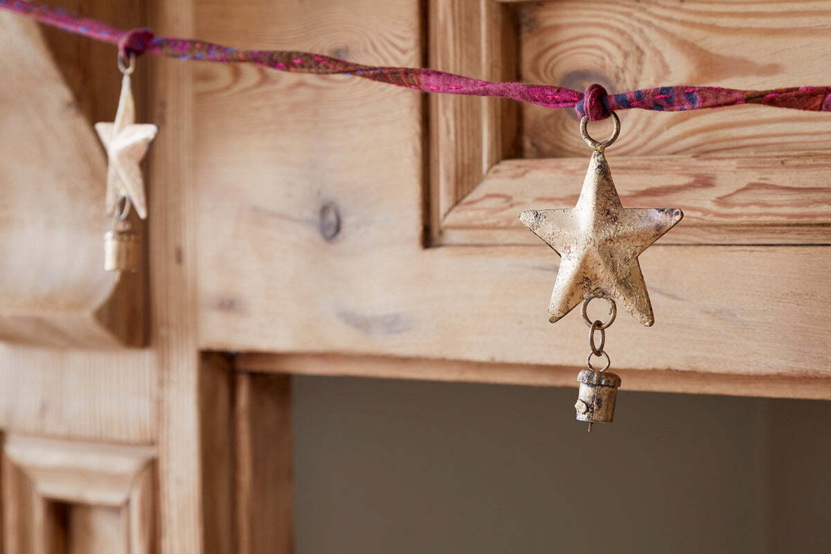 A close-up of the nkuku Didi Star Garland - Antique Brass, featuring antique brass stars and small bells on a twisted pink and red cord, draped over a rustic wooden cabinet with a softly blurred background.