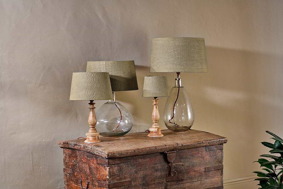 Three Dia Jute Lampshades by nkuku with natural fiber shades and clear glass or wooden bases sit on a rustic wooden chest against a beige wall, complemented by a leafy plant for a cozy, vintage look.