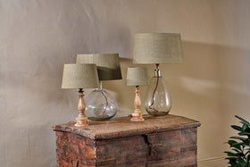 Three nkuku Dia Jute Lampshade table lamps with natural jute shades sit on a rustic chest. Two have clear, textured glass bases; one features a carved wooden base. A green plant appears at the bottom right.