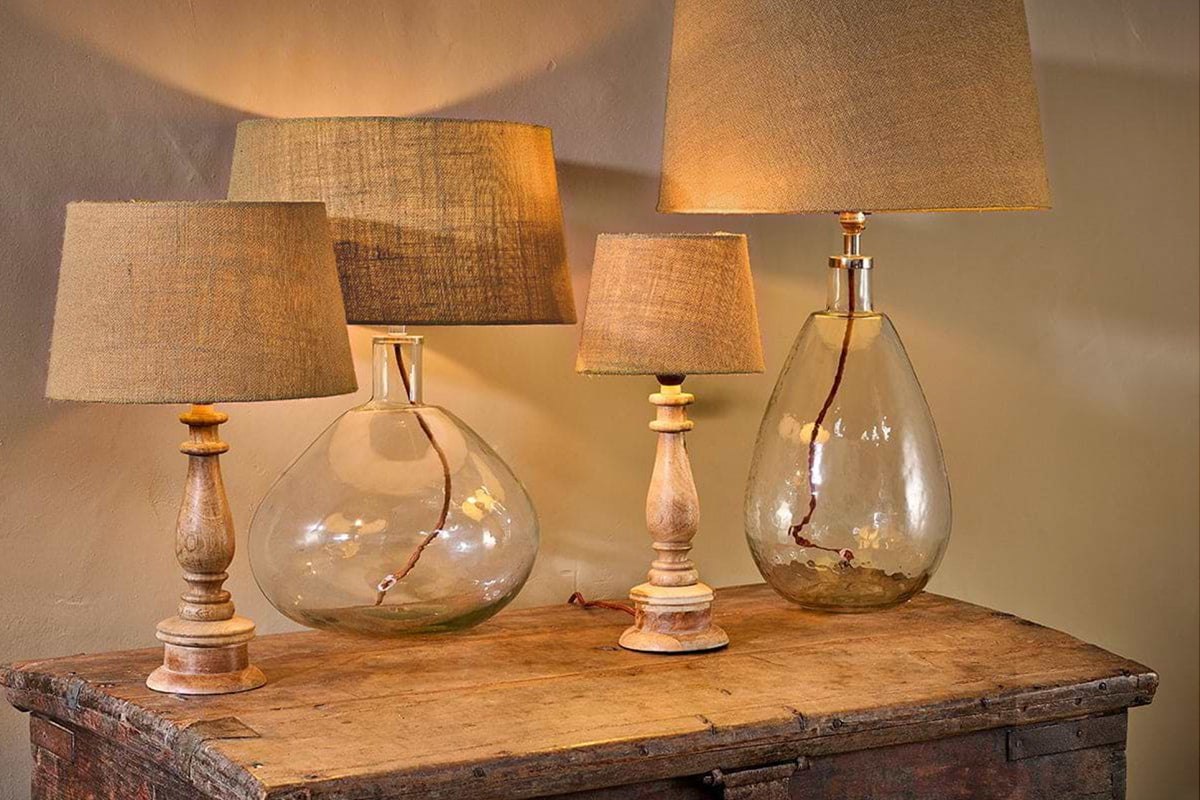 Four nkuku Dia Jute Lampshade - Natural table lamps sit on a rustic wooden chest. Two feature clear glass bases, two have turned wood with a textured finish. Their ambient glow creates a cozy atmosphere against the beige wall.