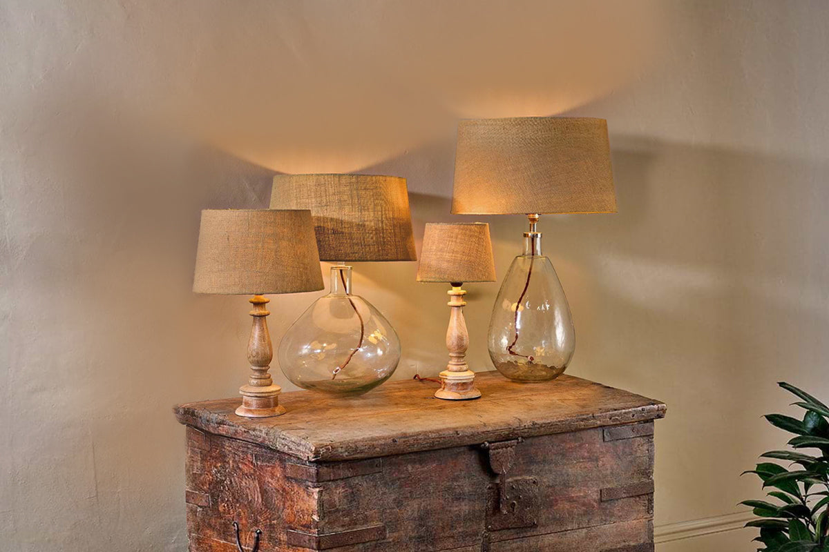 Three nkuku Dia Jute Lampshade table lamps with natural jute shades and glass bases are arranged on a rustic wooden chest. Their varied heights and textured finish are complemented by a leafy green plant beside them.