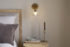 A cozy bedside scene with a wooden headboard, gray pillow, beige bedding, and the nkuku Davika Ribbed Glass Task Wall Lamp in clear and antique brass above a small wooden table holding a bowl, two books, and a brass candle holder with a taper candle.