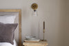 A cozy bedroom features a wooden headboard, grey pillow, white bedding, and a wooden nightstand with decor, all softly illuminated by the nkuku Davika Ribbed Glass Task Wall Lamp in clear & antique brass.