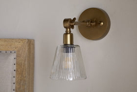 The Davika Ribbed Glass Task Wall Lamp by nkuku, featuring clear ribbed glass and an adjustable antique brass arm, casts a warm glow above a wooden headboard with nailhead trim.