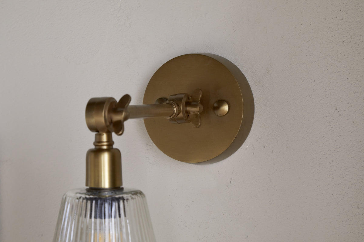 The nkuku Davika Ribbed Glass Task Wall Lamp in clear and antique brass features a round backplate, adjustable arm, and clear ribbed glass shade, shown close-up on a light textured wall.